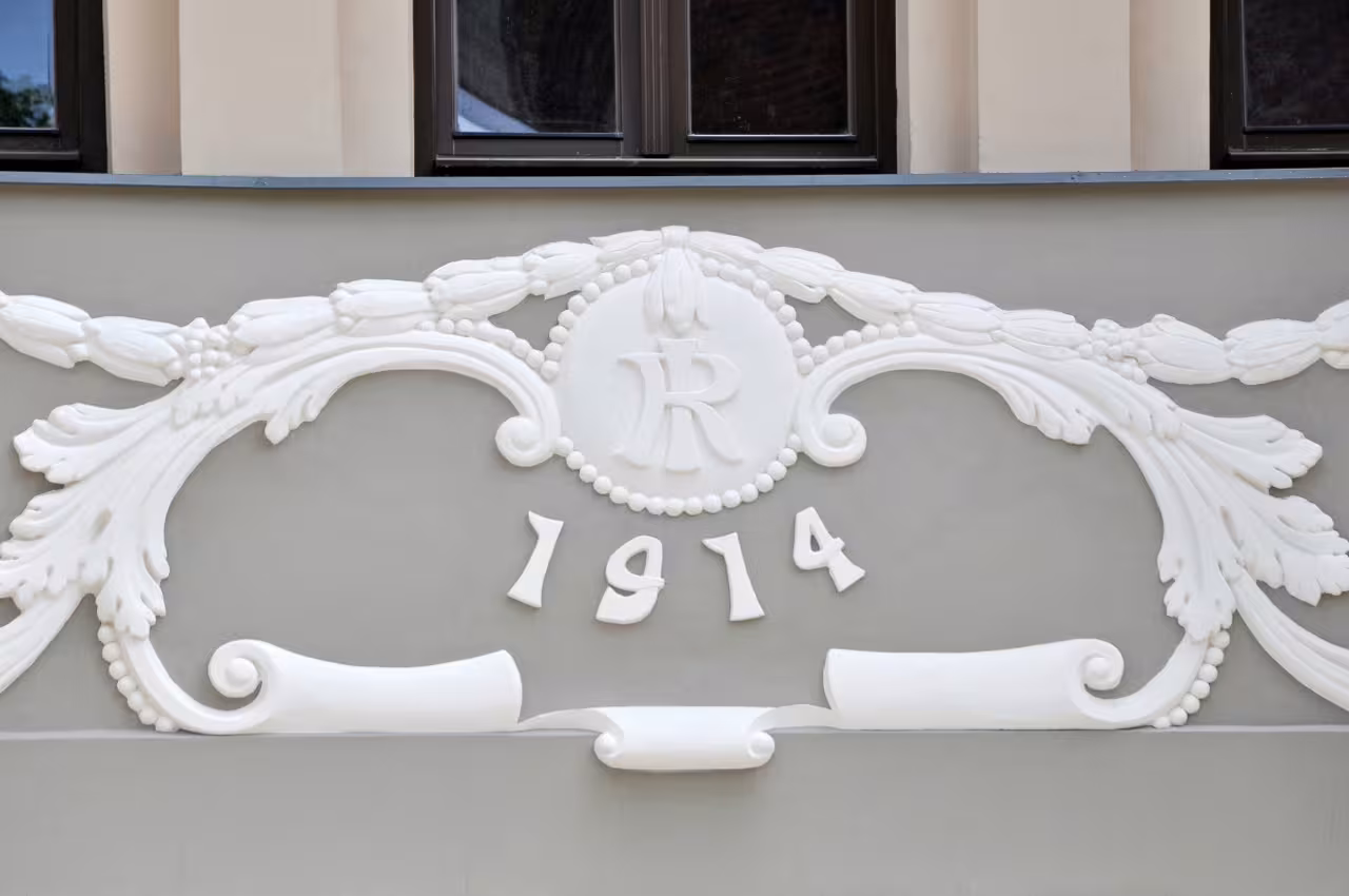 Restored white decorative facade relief displaying the year 1914 and floral motifs on the historical residential building at 5 Alīses Street.