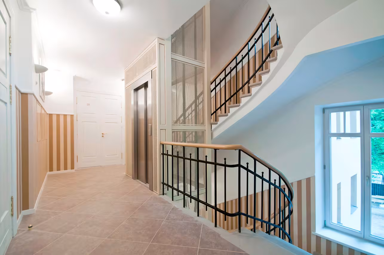 Interior stairwell renovation featuring a modern elevator shaft integrated next to historical railings with striped wallpaper referencing the classical aesthetic.