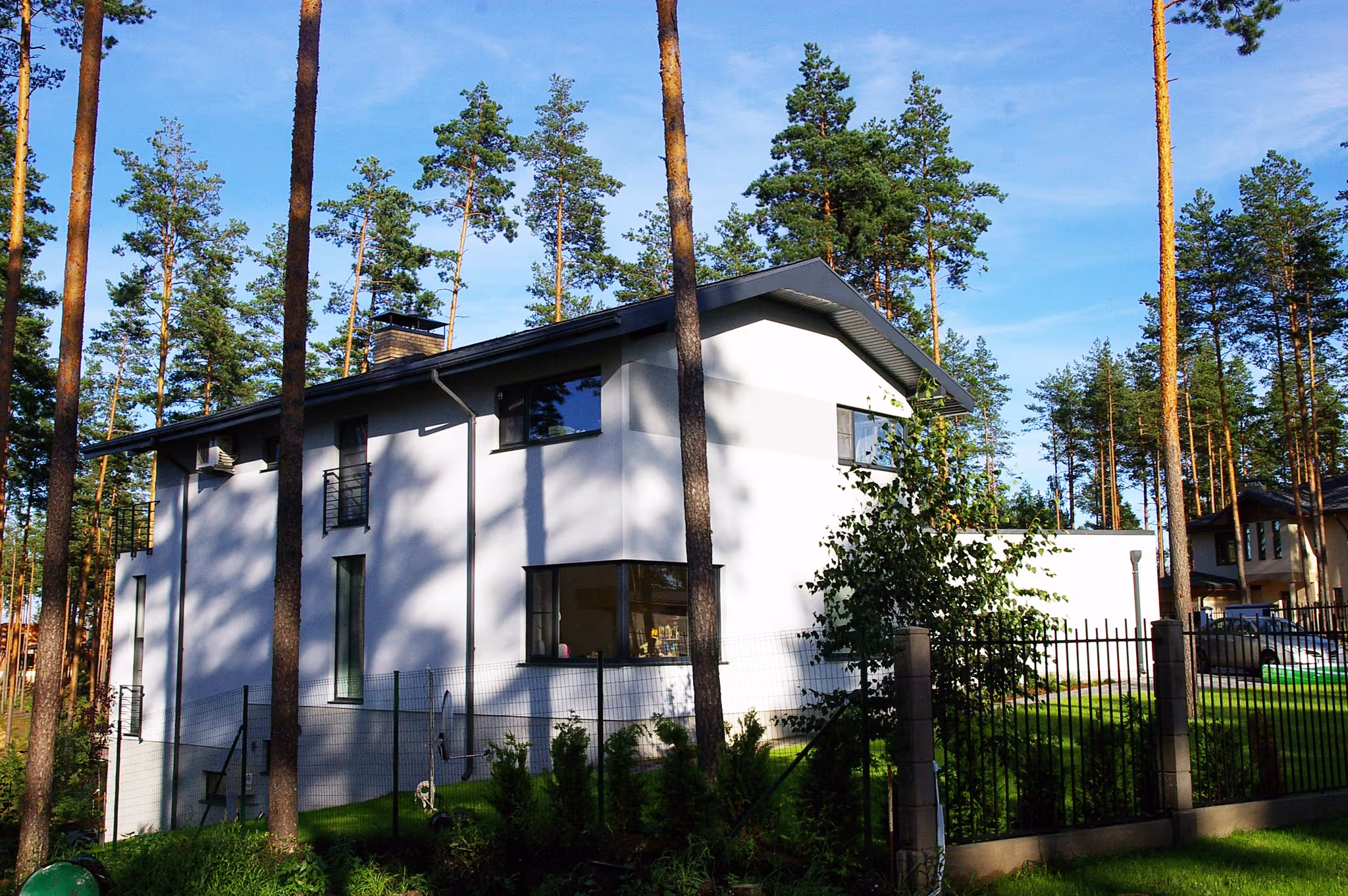 Private house in Ezerkrasti seen behind a metal fence, showing the garage volume, courtyard and integration with surrounding pine trees