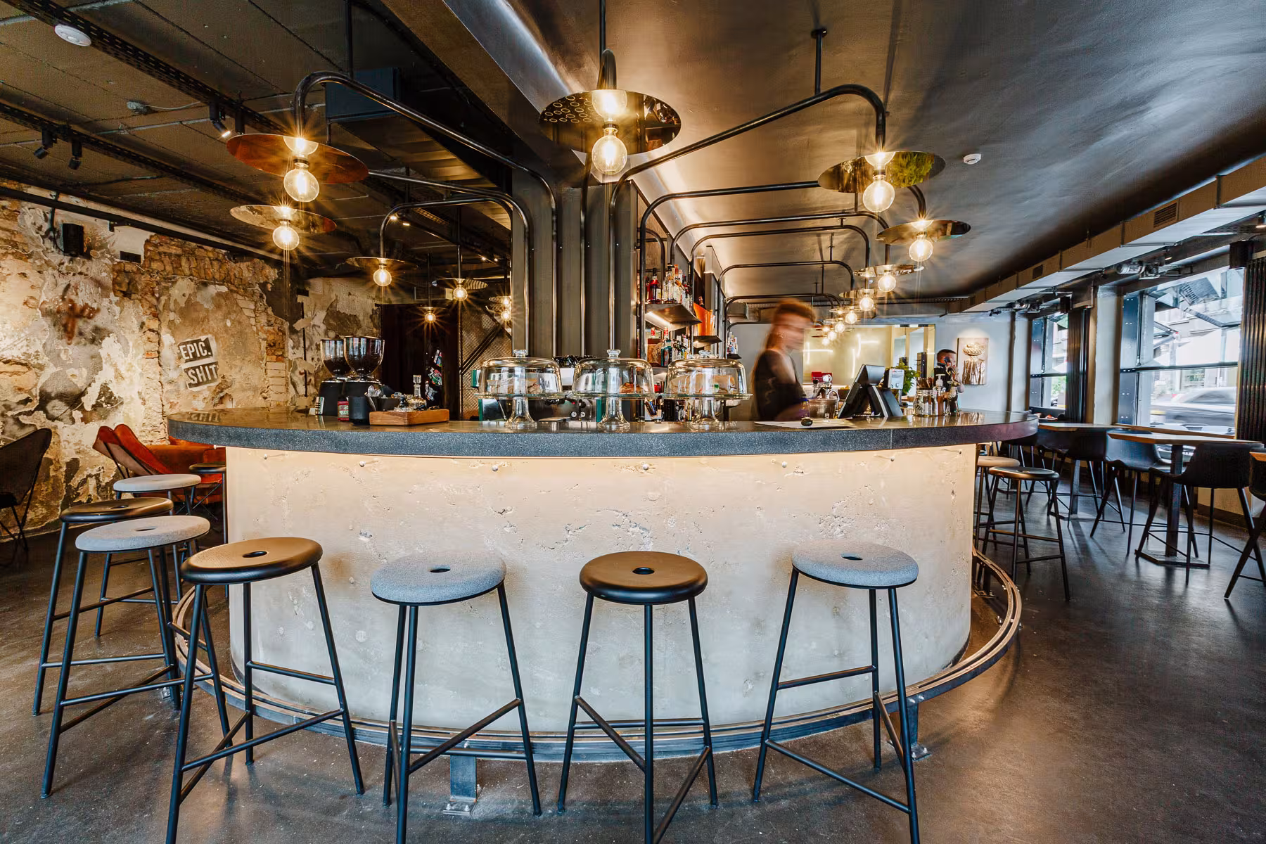 Curved, unpolished concrete bar counter illuminated by a custom structure of black metal pipes and industrial pendant lights.