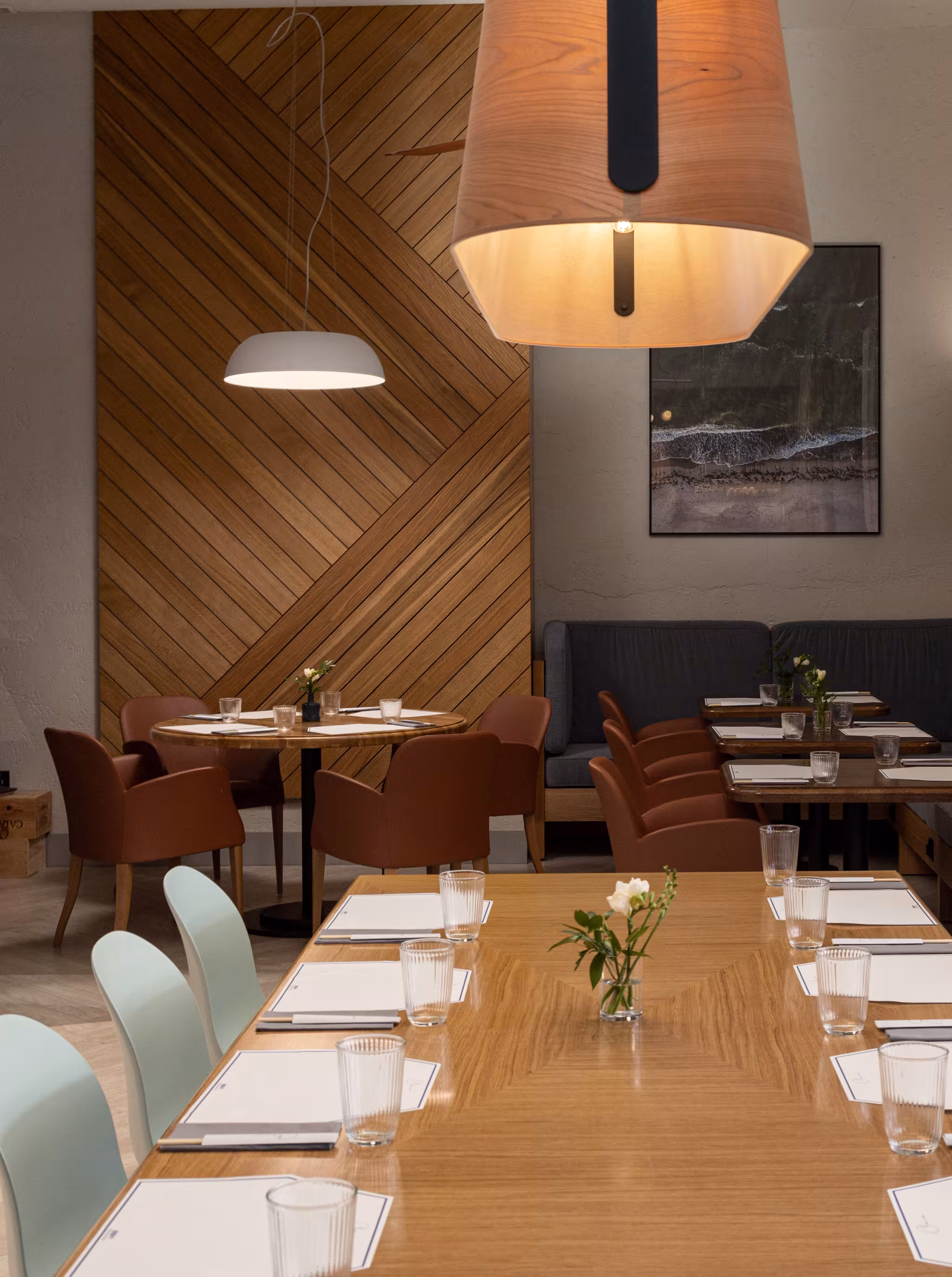 Dining zone illuminated by a custom wooden pendant light, featuring a large-scale aerial beach photograph that connects the interior to the Jūrmala landscape.