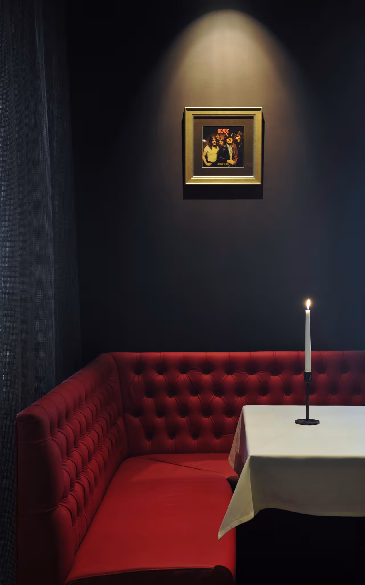 Privāta restorāna sēdvieta ar sarkanu ādas dīvānu, baltu galdautu un izgaismotu AC/DC sienas fotogrāfiju.