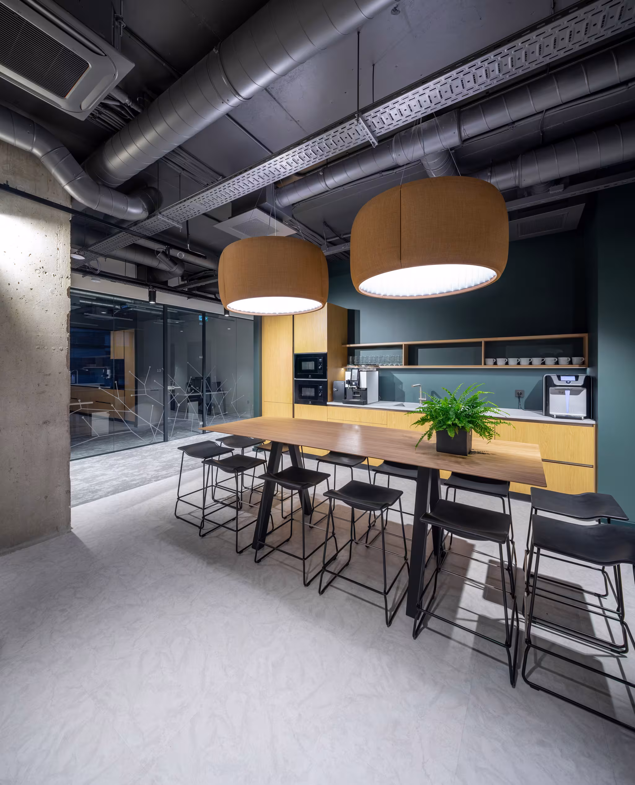 The office kitchen designed as a social hub, featuring a large communal wooden table, oversized acoustic drum pendants and teal cabinetry set against an industrial concrete column.