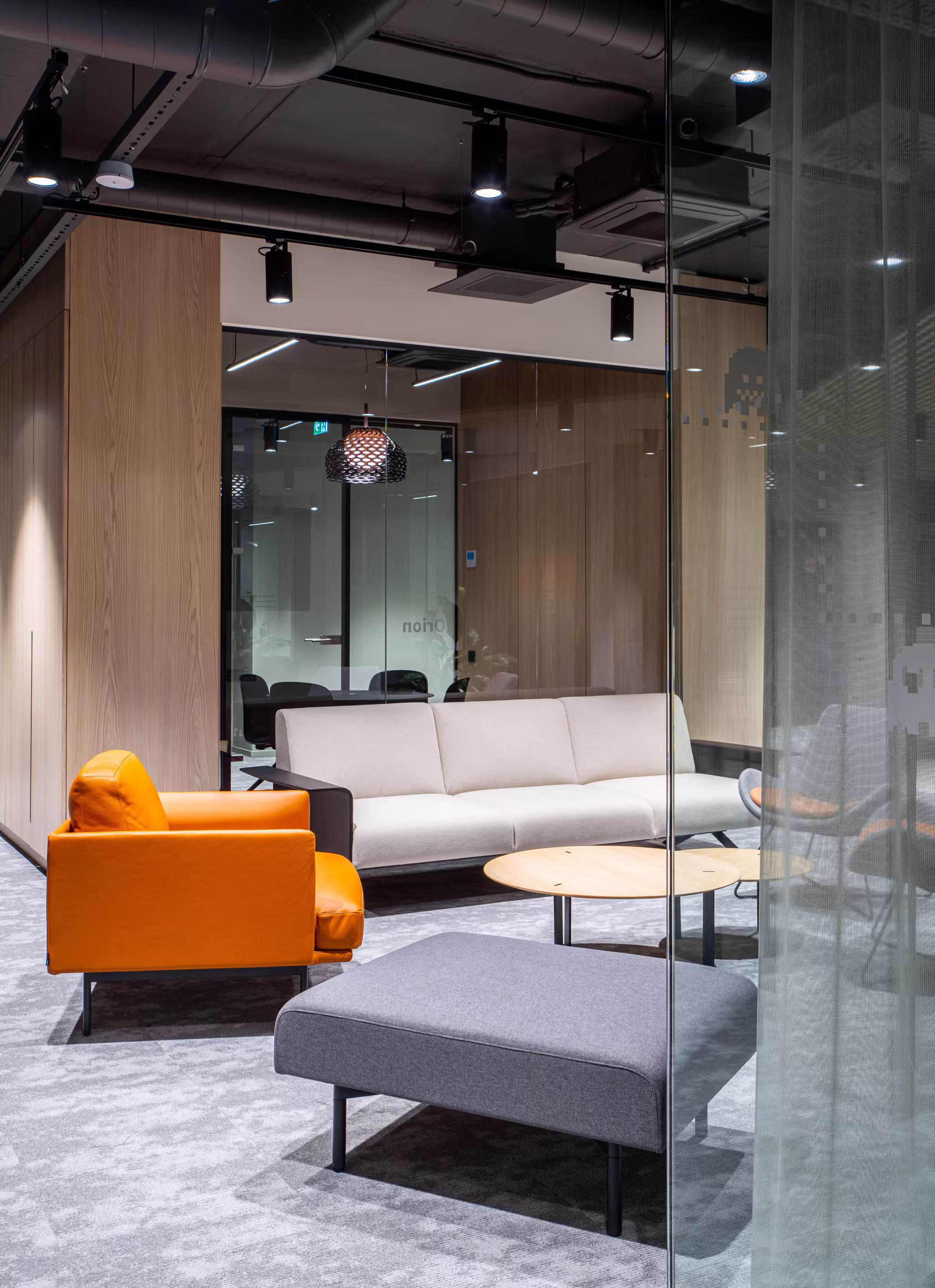 A glimpse into a cozy lounge area through a glass partition, featuring an orange armchair, a light sofa and industrial ceiling details balanced by warm wood tones.
