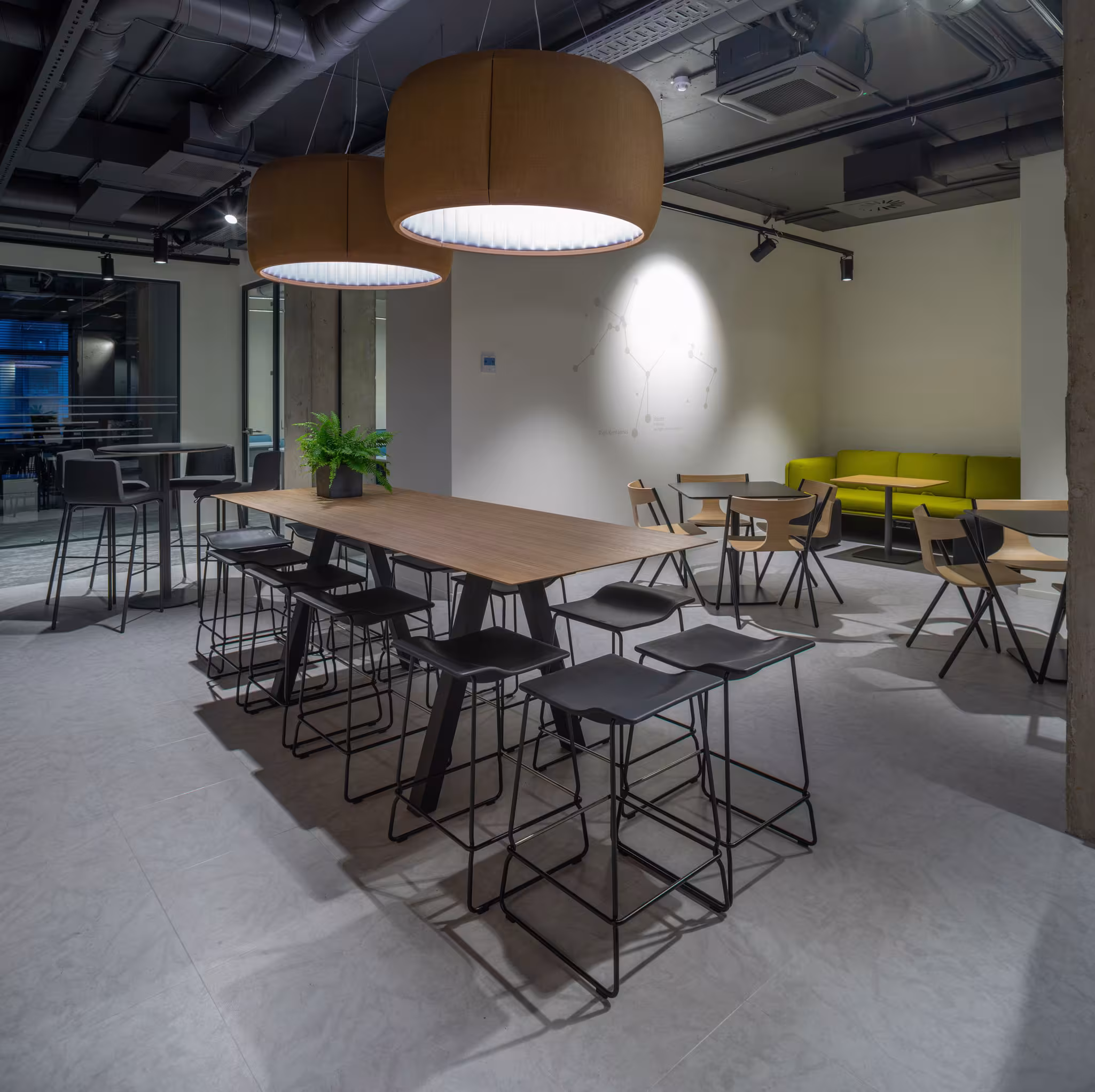 High collaborative table with bar stools and large acoustic drum pendant lights, adjacent to a casual seating area with a green sofa and wall graphics.