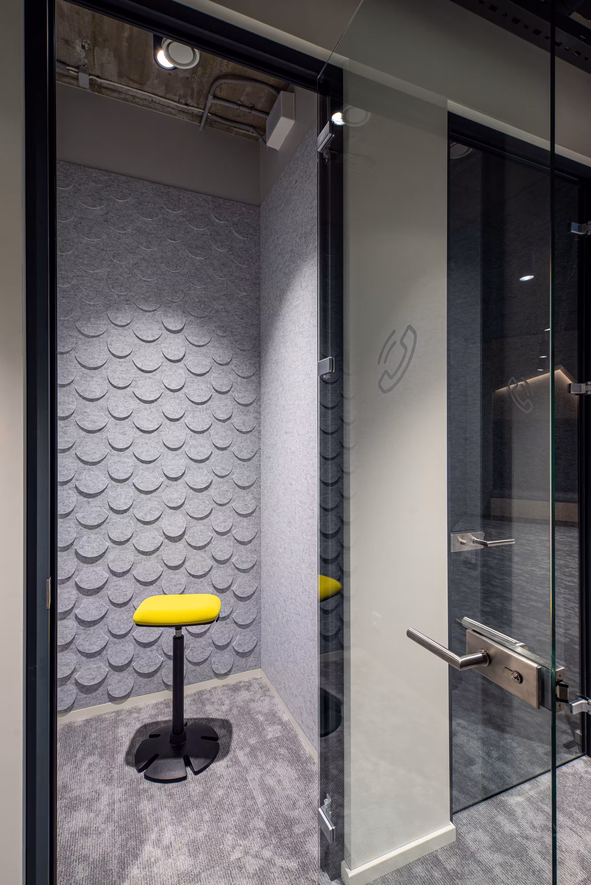 Private phone booth designed for focus, featuring textured grey acoustic wall panels, a yellow stool, and a glass door for transparency.