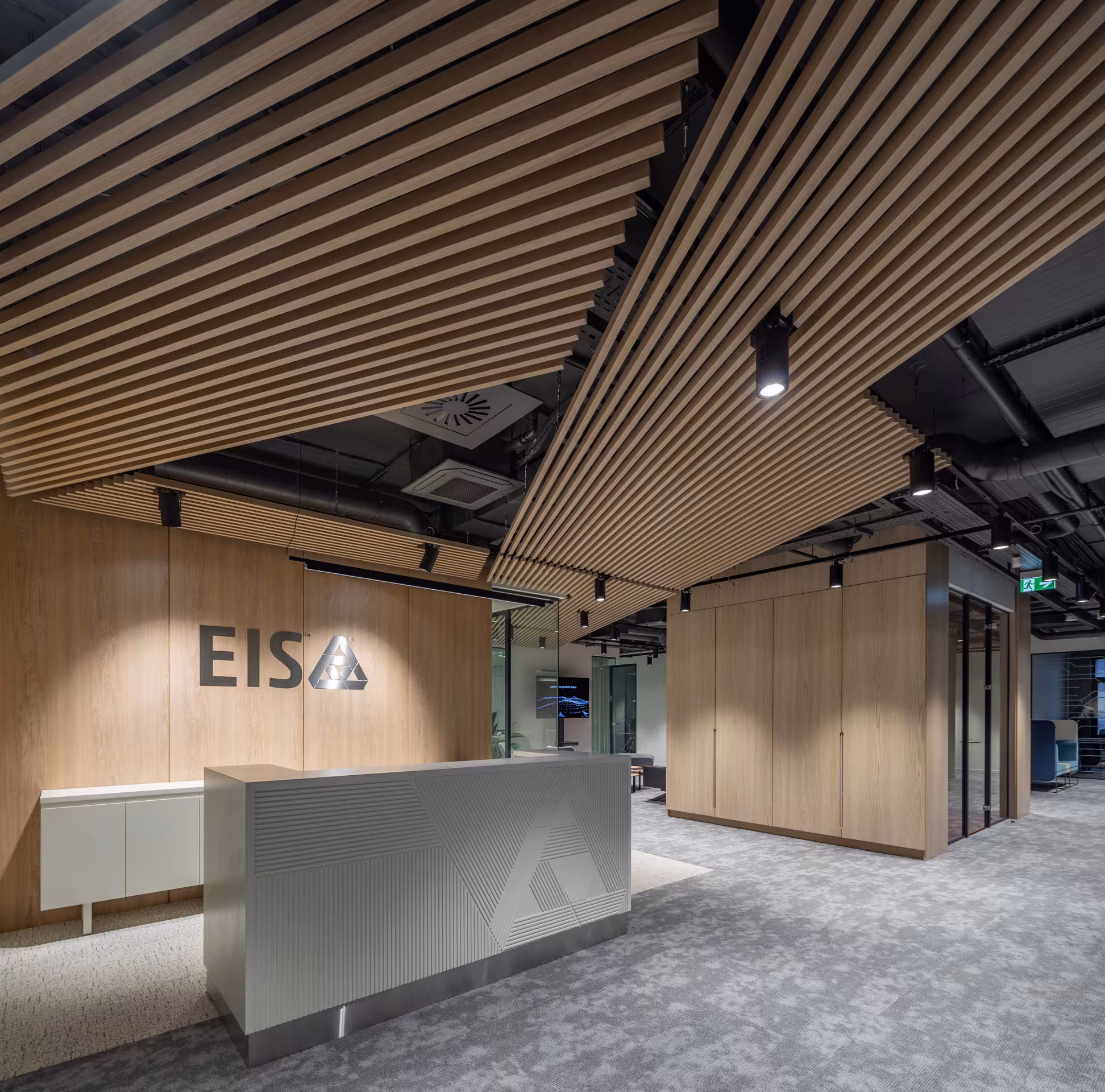 Panoramic view of the reception area highlighting the custom geometric desk, extensive wood paneling and a wooden slat ceiling that balances industrial sleekness with organic warmth.